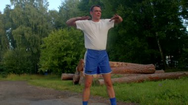 Beyaz gömlekli ve mavi şortlu bir adam parkta güneşli bir günün tadını çıkarır. Kollarını uzatır ve hareketli yaşam tarzını gösterir.