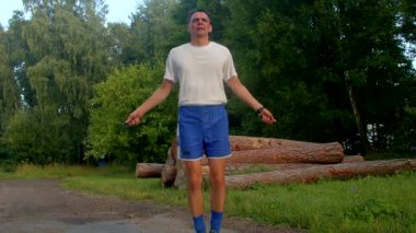 Beyaz gömlekli ve mavi şortlu bir adam parkta, bir kütük tarafından dinlendirilirken, enerjik bir şekilde atlama halatına dönüşüyor, sonra da kendinden emin bir şekilde bir yığın kütük tarafından poz veriyor.