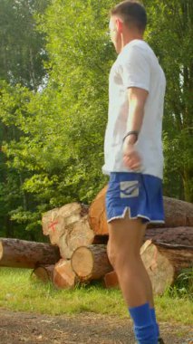 Sakin bir ormanda, beyaz tişörtlü ve mavi şortlu bir adam elinde bir iple bir kütük yığınının önünde duruyor ve telefonuna bakıyor.