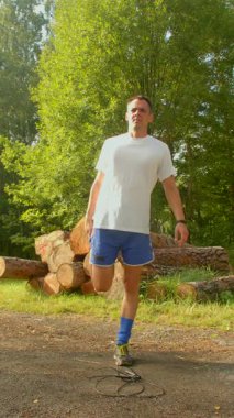 Sakin bir korulukta, beyaz tişörtlü ve mavi şortlu bir adam bacaklarını kırsal bir kütük yığınının yanına uzatarak, kendini adamış olduğunu gösterir.
