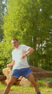 Sakin bir ormanlık alanda, bir adam esnedikten sonra kütüklerin yanında ip atlıyor. Beyaz gömlek ve mavi şort giymiş, sağlık ve etkinliği destekliyor.