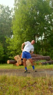 Beyaz tişörtlü ve mavi şortlu bir adam, bir sonraki projesi için potansiyellerine odaklanmış büyük bir odun yığınının yanında güvenle duruyor.