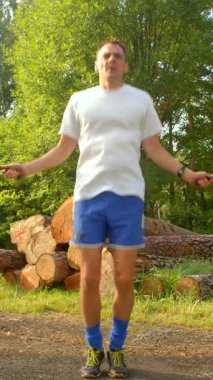 Beyaz gömlekli ve mavi şortlu bir adam çevikliğini bir kütük yığınının önünde ip atlayıp aktif yaşam tarzını vurgulayarak gösteriyor.