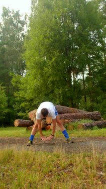Bir adam dingin bir ormandaki engebeli bir odun yığınının yanında duruyor, bacaklarını uzatıyor ve yemyeşil çimlerin arasında doğayla bağ kuruyor.