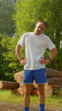 Mavi şortlu, beyaz tişörtlü bir adam yanında büyük bir yığın kütüğün önünde dikiliyor.