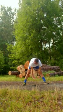 Bu videoda, bir adam kırsal alanda atletizm gösteriyor, amuda kalkarak başlıyor, sonra bacaklarını esnetiyor, ve sopasını kütüklere sallıyor.