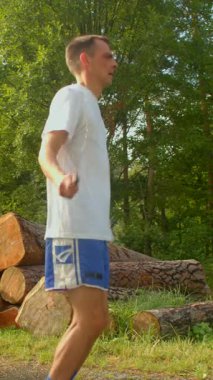 Bir adam sakin bir ormanda koşar, beyaz gömleği ve mavi şortuyla bir kütük yığınının yanında durur, koşusuna devam etmeden önce doğanın tadını çıkarır.