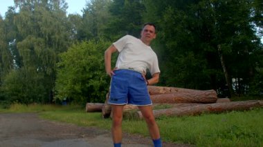 Beyaz tişörtlü ve mavi şortlu bir adam sakin bir parkta esneyerek terledikçe spor yapma kararlılığını ve kararlılığını gösterir.