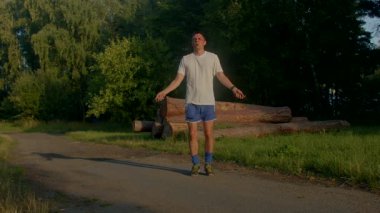 Bir adam halat atlayıp verimli bir ormanda manzaralı bir yolda koşup enerjik pozlar ve açık hava coşkusu yakalayarak fitness rutinini gösterir.