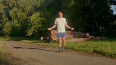 Bir adam güzel açık alanlarda ip atlıyor parklar ve ağaçlık patikalar gibi, zindelik rutinini canlı güzellikler arasında sergiliyor.