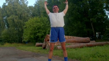 Beyaz gömlekli ve mavi şortlu bir adam parkta egzersiz yapıyor, koşuyor, dikkatli molalar veriyor ve esniyor. Açık havada sergilenen hareketler motivasyona ilham veriyor.