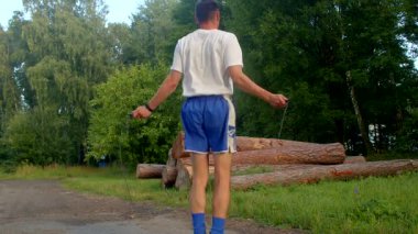 Beyaz gömlekli ve mavi şortlu bir adam canlı bir parkta ip atlıyor, çeviklik sergiliyor, sonra sakince orman yolunda yürüyor.