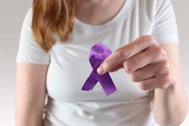 Genç beyaz kadın elinde mor bir farkındalık kurdelesi tutuyor Ev İçi Şiddet ve Alzheimer Bilinci.