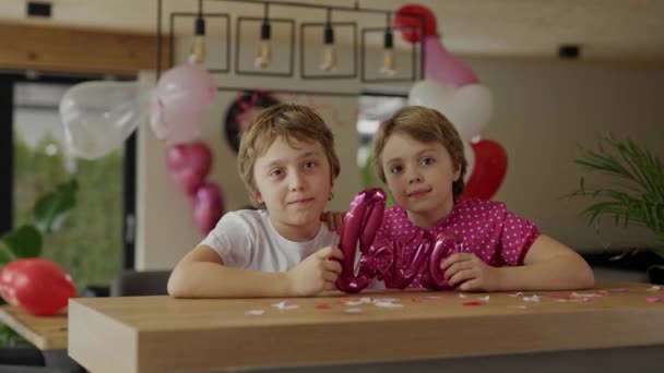 男の子と女の子はバレンタインデーの装飾された部屋のバーテーブルに座っています — ストック動画
