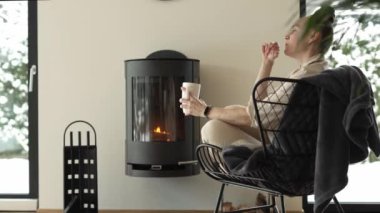 Female sits in the armchair close to the real chimney, drinks cup of hot tea and speaks to someone. High quality 4k footage