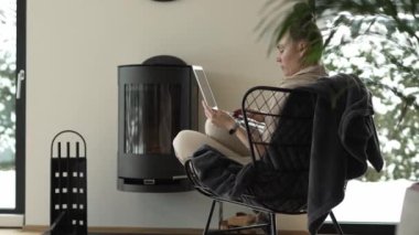 Female sits in the armchair close to the real chimney and works at laptop. High quality 4k footage