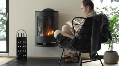 Female sits in the armchair close to the real chimney and reads a book. High quality 4k footage