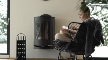 Female sits in the armchair close to the real chimney and reads a book. High quality 4k footage