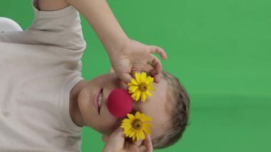 Face of funny boy of 9 years with red nose and yellow daisies. Green screen background. Closeup. High quality footage