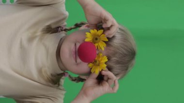 Face of funny girl of 7 years with red nose and yellow daisies. Green screen background. Closeup. High quality footage