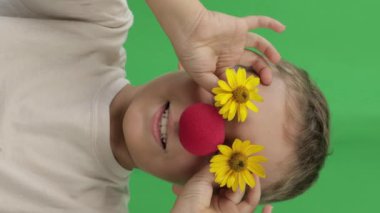 Face of funny boy of 9 years with red nose and yellow daisies. Green screen background. Closeup. High quality footage