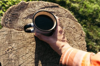 Bir adam elinde bir fincan kahve tutuyor, arka planda yeşil çimenler ve yaşlı bir ağaç var..