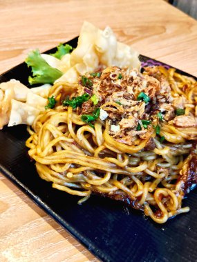 Kızarmış soğanla kaplı Endonezya usulü kızarmış şehriye ve ahşap bir masada siyah bir tabakta gevrek wonton ile servis ediliyor..