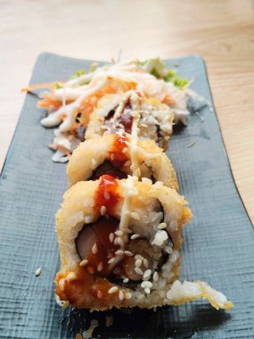 Crispy deep-fried sushi rolls topped with spicy mayo and toasted sesame seeds on a rectangular, textured black plate. The sushi and salad are placed on a light wooden table, enhancing the overall presentation.