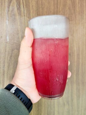 Koyu kırmızı bir içecekle dolu kaburgalı bir bardağı tutan bir el, muhtemelen kuşüzümü suyu, buz küpleriyle birleşmiş. Camın dışı buğulanmış, bu da soğuk olduğunu gösteriyor. Kişi yeşil bir kol ve siyah bir saat takıyor.. 