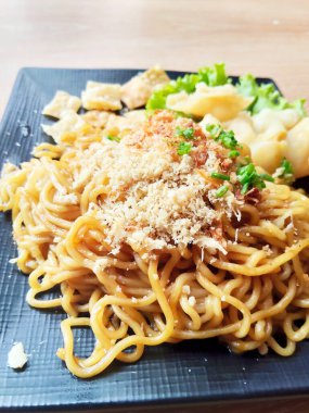 Lezzetli bir porsiyon lezzetli Endonezyalı Mie Goreng (kızarmış erişte) siyah kare tabakta, kızarmış hamur tatlısı ve bol miktarda kızarmış soğan ile servis edilir.