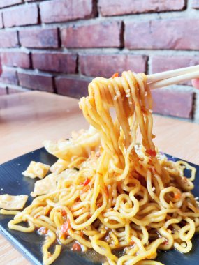 Lezzetli bir porsiyon lezzetli Endonezyalı Mie Goreng (kızarmış erişte) siyah kare tabakta, kızarmış hamur tatlısı ve bol miktarda kızarmış soğan ile servis edilir.