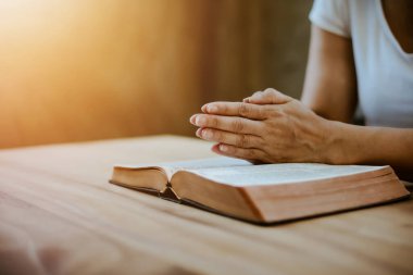 Bir kadının elleri kapalı bir masada açık kutsal incilin üzerinde dua ederken pencereleri açık ve sıcak bir tonda uzanır. Kopya alanı ile Hristiyan inancı ve güven kavramı. Hıristiyan bağlılık geçmişi.