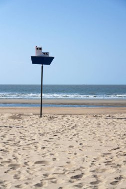 Wooden model of boat on pole on sandy beach with real big cargo ship in background, sunny day