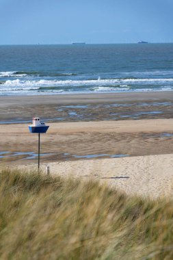Wooden model of boat on pole on sandy beach with real big cargo ship in background, sunny day