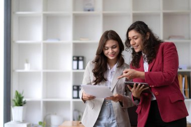 Two beautiful Asian businesswomen standing using digital tablet consulting and analyzing information in office work...