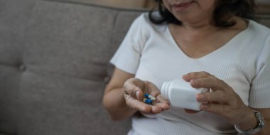 Yaşlı Güneydoğu Asyalı kadın elinde kalp hastalığı hapları tutarken koltukta tek başına oturuyor, yaşlı sağlık kavramı..