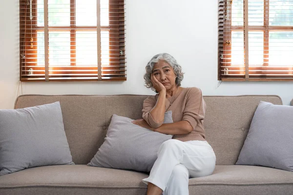 Thoughtful mature woman sitting on couch at home alone, dreaming and planning, serious pensive older senior female looking into distance, in window in living room.