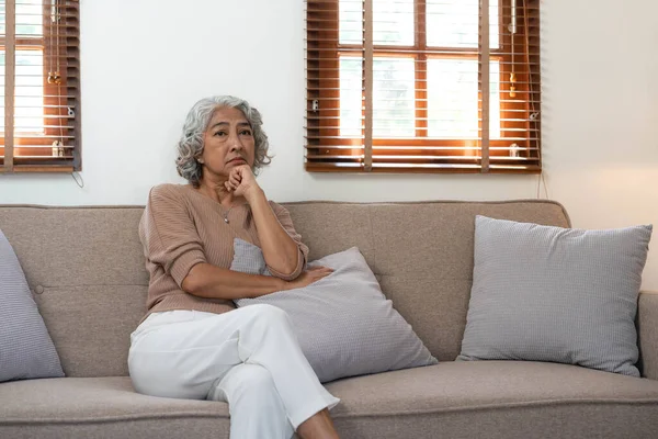 Thoughtful mature woman sitting on couch at home alone, dreaming and planning, serious pensive older senior female looking into distance, in window in living room.