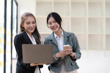 Two beautiful Asian businesswomen standing using digital tablet consulting and analyzing information in office work...