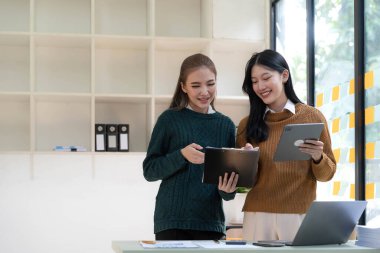 Two beautiful Asian businesswomen standing using digital tablet consulting and analyzing information in office work...