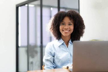 Ev ofisinde online ile bir dizüstü bilgisayar çalışma masasında oturan genç Afrikalı kadın girişimci gülümseyen.