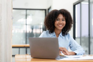 Ev ofisinde online ile bir dizüstü bilgisayar çalışma masasında oturan genç Afrikalı kadın girişimci gülümseyen.
