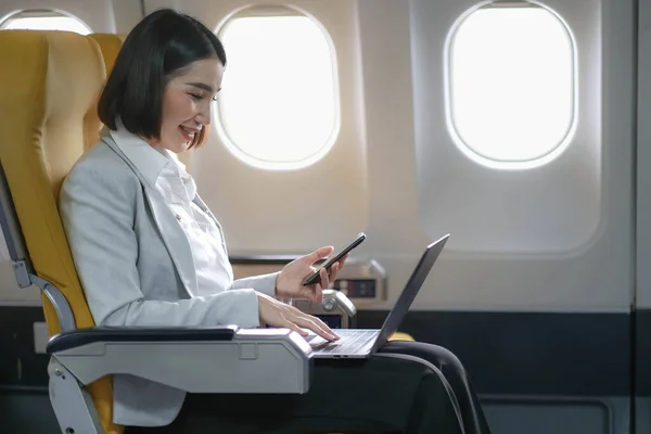 Asian woman passenger sitting in airplane near window and reading news from social networks or using travel applications in smartphone.