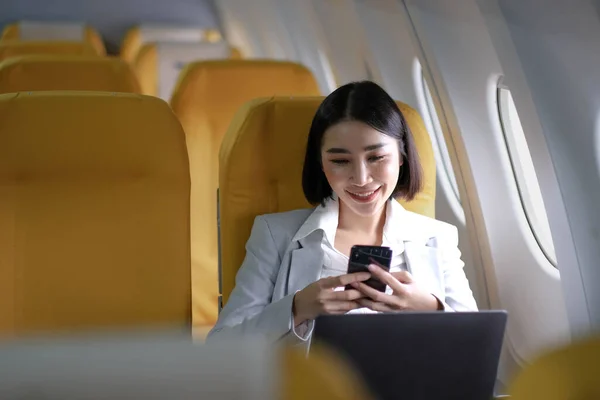 Asian woman passenger sitting in airplane near window and reading news from social networks or using travel applications in smartphone.