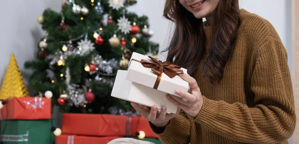 Smiling asian woman in festive mood holding wrapped gift box with brown ribbon in hands, stretching out present to camera, celebrating Merry Christmas
