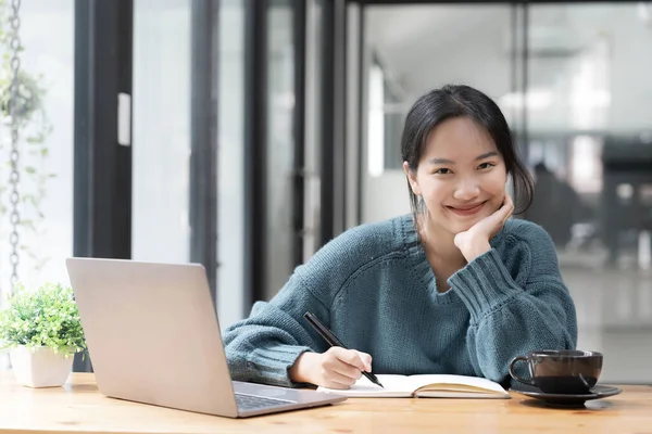 Beautiful women student studying online takes notes on her laptop to gather information about her work smiling face and a happy study posture