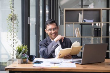 Takım elbiseli, elinde sıcak kahve fincanı olan genç Asyalı iş adamı ofisteki dizüstü bilgisayarına bakıyor. Takım elbiseli adam iyi aydınlatılmış iş yerinde dizüstü bilgisayar kullanıyor....