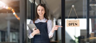 Portrait of a happy waitress standing at restaurant entrance. Portrait of mature business womanattend new customers in her coffee shop. Happy woman owner showing open sign in her small business shop
