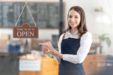 Portrait of a happy waitress standing at restaurant entrance. Portrait of mature business womanattend new customers in her coffee shop. Happy woman owner showing open sign in her small business shop