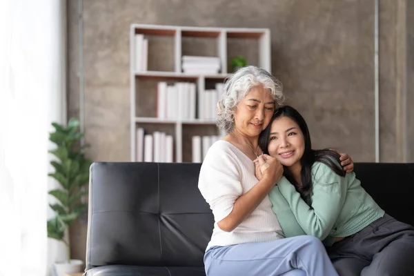 Loving adult daughter hugging older mother, standing behind couch at home, family enjoying tender moment together, young woman and mature mum or grandmother looking at each other, two generations..